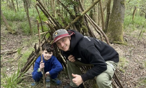 Adult and child in front of a wooden den built of sticks at Salcey Forest